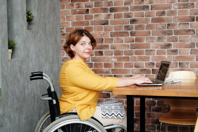 A woman in a yellow sweater in a wheelchair, typing on a laptop.