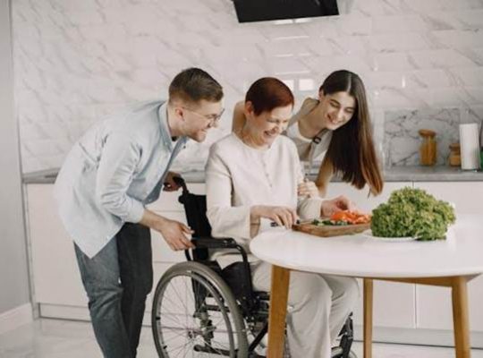 Family supporting a woman in a wheelchair while cooking in a kitchen, showing the importance of customising temporary housing during a move for accessibility and comfort. https://www.pexels.com/photo/women-and-man-with-vegetables-on-table-in-kitchen-7446746/