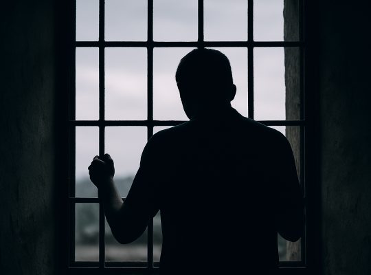 Image credit: © OpenAI / Generated with DALL·E — “Silhouette of a person behind bars symbolising detention and loss of freedom.