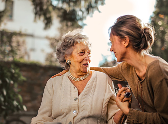 Image Description: Two women sitting together with the younger woman facing the elderly woman and her arm stretched out behind the elderly woman's Back. Image Credit: https://www.pexels.com/photo/3768131/