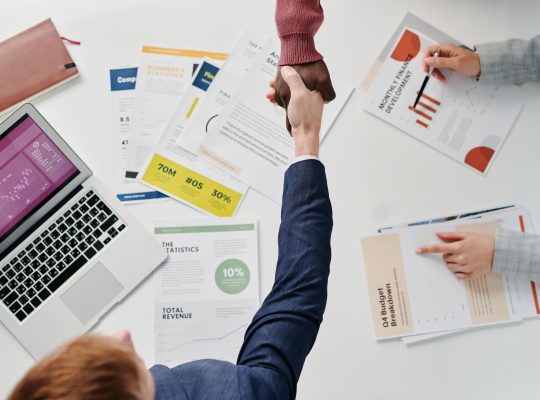 Image Description: People doing a handshake at work, Three business people working at a table. Two of the business people are reaching across the table to shake hands. Image Credit: Tiger Lily - https://www.pexels.com/@tiger-lily/ Image Source: https://www.pexels.com/photo/people-doing-hand-shake-at-work-7109292