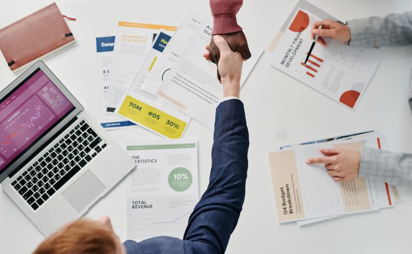 Image Description: People doing a handshake at work, Three business people working at a table. Two of the business people are reaching across the table to shake hands. Image Credit: Tiger Lily - https://www.pexels.com/@tiger-lily/ Image Source: https://www.pexels.com/photo/people-doing-hand-shake-at-work-7109292